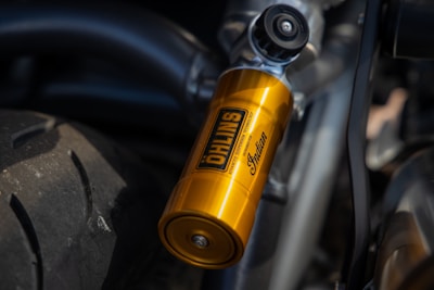 A gold-colored Öhlins advanced suspension technology component is prominently displayed, mounted on a vehicle. Nearby, part of a black tire is visible, along with metallic components that suggest the suspension system of a motorcycle or similar machinery. The setting appears rugged, possibly outdoors, evidenced by the lighting and dust on the tire.