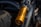 A gold-colored Öhlins advanced suspension technology component is prominently displayed, mounted on a vehicle. Nearby, part of a black tire is visible, along with metallic components that suggest the suspension system of a motorcycle or similar machinery. The setting appears rugged, possibly outdoors, evidenced by the lighting and dust on the tire.