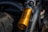 A gold-colored Öhlins advanced suspension technology component is prominently displayed, mounted on a vehicle. Nearby, part of a black tire is visible, along with metallic components that suggest the suspension system of a motorcycle or similar machinery. The setting appears rugged, possibly outdoors, evidenced by the lighting and dust on the tire.
