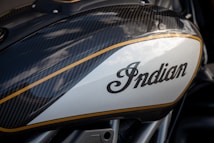 Close-up of a motorcycle fuel tank with a carbon fiber texture and a large logo with the word 'Indian' in a stylish script. The tank features sleek, aerodynamic design elements accented with a gold stripe along the edges, reflecting a modern and high-performance aesthetic.