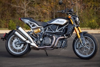 A sleek motorcycle is positioned on a paved surface with a wooded background. The bike features a modern design with polished metal components and a prominent exhaust system. The frame showcases intricate engineering details, and the bike's brand logo is visible on the fuel tank.