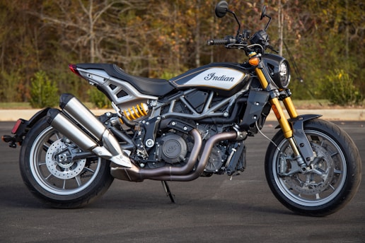 A sleek motorcycle is positioned on a paved surface with a wooded background. The bike features a modern design with polished metal components and a prominent exhaust system. The frame showcases intricate engineering details, and the bike's brand logo is visible on the fuel tank.