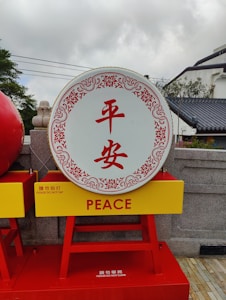 A large decorative drum with the Chinese characters '平安' in the center, surrounded by an ornate red border. Below the drum, there is a yellow sign with red text reading 'PEACE' and additional instructions 'PLEASE DO NOT TAP'. The drum is mounted on a red stand. The background includes a gray stone wall with some greenery and a traditional style building with a tiled roof.