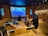A room with wooden flooring and walls featuring a large screen displaying a serene river scene. Three children are seated on the floor, facing the screen alongside sculptures of a bear and a cat. The atmosphere appears calm and contemplative, with natural-themed decor on the walls.