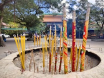 Large incense sticks, adorned with colorful patterns, are burning in a circular stone holder filled with ash. The setting is outdoors, with tall trees providing shade and a traditional building with a tiled roof in the background.