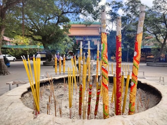 Large incense sticks, adorned with colorful patterns, are burning in a circular stone holder filled with ash. The setting is outdoors, with tall trees providing shade and a traditional building with a tiled roof in the background.