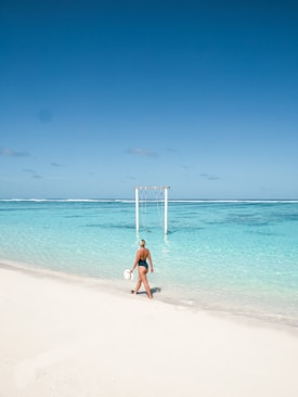 A person wearing a swimsuit walks along a pristine white sandy beach, carrying a sun hat in hand. The turquoise ocean stretches out in the background, with a clear blue sky above. A wooden swing set is partially submerged in the shallow water.