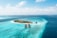 Aerial view of an idyllic tropical island surrounded by clear turquoise waters. The island is lush with green palm trees and vegetation. Several overwater bungalows extend into the shallow blue lagoon, creating a scenic and luxurious resort setting. The deep blue ocean surrounds the bright sandy beaches and vibrant coral reefs.