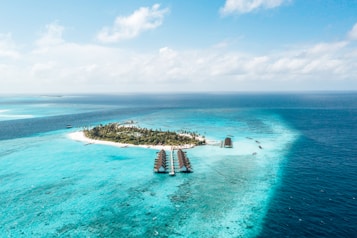 Aerial view of an idyllic tropical island surrounded by clear turquoise waters. The island is lush with green palm trees and vegetation. Several overwater bungalows extend into the shallow blue lagoon, creating a scenic and luxurious resort setting. The deep blue ocean surrounds the bright sandy beaches and vibrant coral reefs.