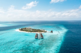 Aerial view of an idyllic tropical island surrounded by clear turquoise waters. The island is lush with green palm trees and vegetation. Several overwater bungalows extend into the shallow blue lagoon, creating a scenic and luxurious resort setting. The deep blue ocean surrounds the bright sandy beaches and vibrant coral reefs.