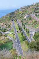 An expansive view of a lush, green landscape featuring a modern highway with multiple lanes and well-maintained roads. Two long tunnels are visible, situated in a hilly area with scattered residential houses. The background shows the ocean under a bright, clear sky, adding to the scenic beauty of the area.