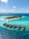Aerial view of a tropical island resort with overwater bungalows connected by a wooden walkway. The turquoise water surrounds the island, creating a vibrant contrast with the lush greenery and sandy beaches.