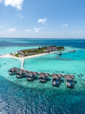 Aerial view of a tropical island resort with overwater bungalows connected by a wooden walkway. The turquoise water surrounds the island, creating a vibrant contrast with the lush greenery and sandy beaches.