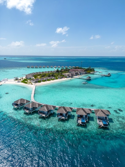 Aerial view of a tropical island resort with overwater bungalows connected by a wooden walkway. The turquoise water surrounds the island, creating a vibrant contrast with the lush greenery and sandy beaches.