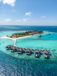 Aerial view of a tropical island resort with overwater bungalows connected by a wooden walkway. The turquoise water surrounds the island, creating a vibrant contrast with the lush greenery and sandy beaches.