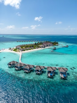 Aerial view of a tropical island resort with overwater bungalows connected by a wooden walkway. The turquoise water surrounds the island, creating a vibrant contrast with the lush greenery and sandy beaches.