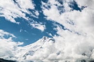 A majestic mountain peak piercing through fluffy white clouds under a blue sky.