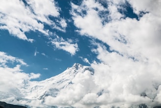 A majestic mountain peak piercing through fluffy white clouds under a blue sky.