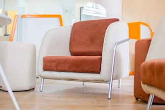 A modern chair with a combination of beige and rust-orange upholstery sits on a wooden floor. The chair features metallic legs and is part of a contemporary room setting. Other similar chairs and a small round table are visible in the background, along with an orange and white room decor.