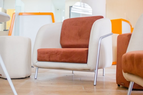 A modern chair with a combination of beige and rust-orange upholstery sits on a wooden floor. The chair features metallic legs and is part of a contemporary room setting. Other similar chairs and a small round table are visible in the background, along with an orange and white room decor.