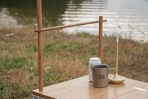 A tranquil outdoor setting with a wooden table holding a can, a mug with a logo, and a candle holder. The background features a calm body of water and grass, suggesting a lakeside or riverside location.