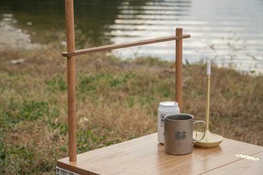 A tranquil outdoor setting with a wooden table holding a can, a mug with a logo, and a candle holder. The background features a calm body of water and grass, suggesting a lakeside or riverside location.