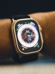 A close-up view of a smartwatch on a person's wrist. The watch displays a colorful digital clock face with various indicators such as heart rate and activity rings. The strap is black, and the watch body is metallic.