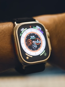A close-up view of a smartwatch on a person's wrist. The watch displays a colorful digital clock face with various indicators such as heart rate and activity rings. The strap is black, and the watch body is metallic.