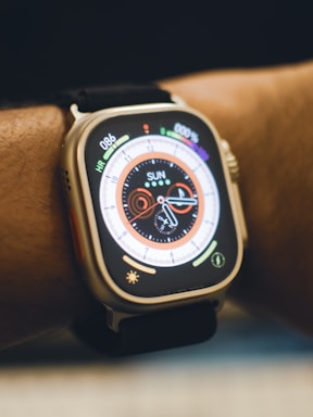 A close-up view of a smartwatch on a person's wrist. The watch displays a colorful digital clock face with various indicators such as heart rate and activity rings. The strap is black, and the watch body is metallic.