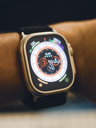 A close-up view of a smartwatch on a person's wrist. The watch displays a colorful digital clock face with various indicators such as heart rate and activity rings. The strap is black, and the watch body is metallic.
