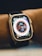 A close-up view of a smartwatch on a person's wrist. The watch displays a colorful digital clock face with various indicators such as heart rate and activity rings. The strap is black, and the watch body is metallic.