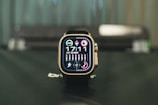 A smartwatch with a square face sitting on a surface, displaying the time 17:11 with various icons and colorful rings indicating activity and notifications. The watch has a modern design with a metallic frame and is positioned against a blurred background.