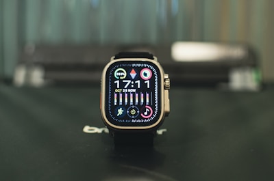 A smartwatch with a square face sitting on a surface, displaying the time 17:11 with various icons and colorful rings indicating activity and notifications. The watch has a modern design with a metallic frame and is positioned against a blurred background.