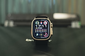 A smartwatch with a square face sitting on a surface, displaying the time 17:11 with various icons and colorful rings indicating activity and notifications. The watch has a modern design with a metallic frame and is positioned against a blurred background.