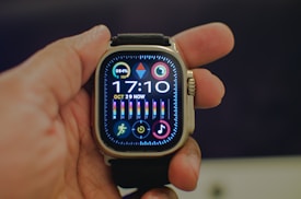 A hand holding a smartwatch with a black strap. The smartwatch screen displays various colorful icons and information such as the time (17:10), the date (Oct 29), and additional icons for fitness tracking, music, and other applications. The battery level indicator is at 84%. The screen is vibrant with multiple colors and detailed graphics.