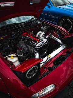 A close-up view of a car engine with the hood open, displaying components like pipes and a polished metal cover. The car's exterior is red, and another blue car is parked nearby, partially visible.