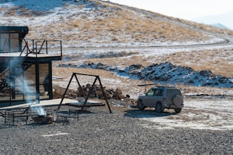 A modern cabin with large glass windows is situated in a winter landscape with patches of snow and dry grass. A fire pit with smoke rising and outdoor seating is visible in the foreground. A small SUV is parked nearby, with a pile of firewood next to it. The terrain is hilly with a winding path in the background.