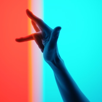 A hand is silhouetted against a split background, with one side in red and the other in cyan. The hand is positioned with fingers spread apart, creating a striking contrast between the two vibrant colors.