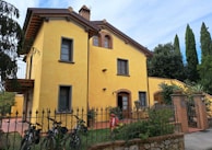 A charming, two-story yellow house with a tiled roof and arched windows, surrounded by greenery and a wrought-iron fence. Three bicycles are parked near the entrance, and there are tall trees and a clear blue sky in the background.