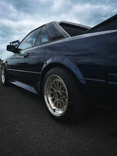 A classic sports car featuring a sleek, angular design and reflective dark paint. The vehicle has stylish, gold multi-spoke wheels, and is positioned on a dark asphalt surface. The background shows an overcast sky, creating a moody atmosphere.