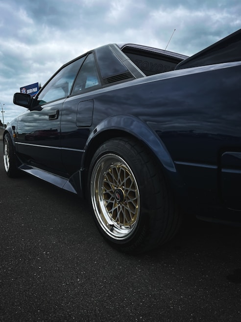 A classic sports car featuring a sleek, angular design and reflective dark paint. The vehicle has stylish, gold multi-spoke wheels, and is positioned on a dark asphalt surface. The background shows an overcast sky, creating a moody atmosphere.