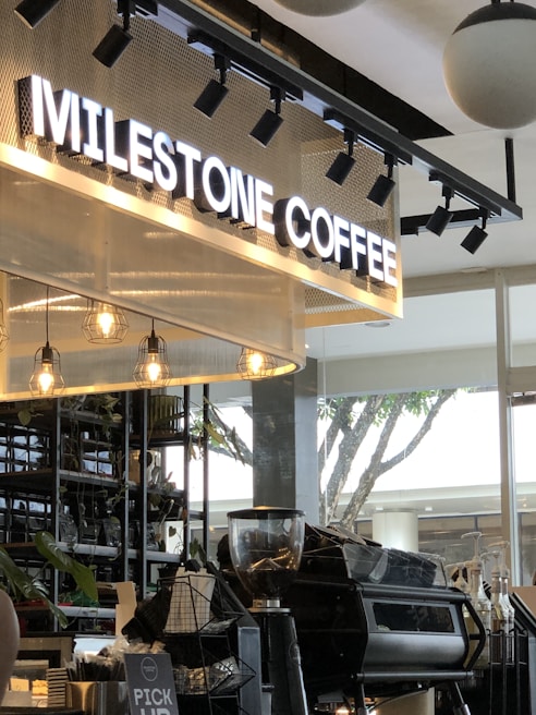 A modern coffee shop setting featuring a large illuminated sign that reads 'Milestone Coffee.' The interior includes hanging pendant lights, a coffee counter with a grinder and espresso machine, and decorative plants on shelves. Large windows allow natural light to flood in, while a tree can be seen outside.