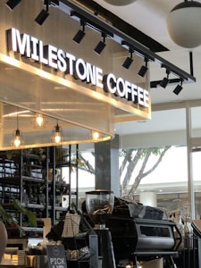 A modern coffee shop setting featuring a large illuminated sign that reads 'Milestone Coffee.' The interior includes hanging pendant lights, a coffee counter with a grinder and espresso machine, and decorative plants on shelves. Large windows allow natural light to flood in, while a tree can be seen outside.
