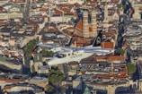 An aircraft labeled 'Alpen Air' flies over a densely packed urban area characterized by historical architecture. Prominent buildings with red-tiled roofs stand out, including notable cathedrals with large domed towers. The city streets are lined with trees and various styles of architectural structures, reflecting a blend of modern and traditional designs.