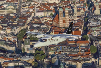 An aircraft labeled 'Alpen Air' flies over a densely packed urban area characterized by historical architecture. Prominent buildings with red-tiled roofs stand out, including notable cathedrals with large domed towers. The city streets are lined with trees and various styles of architectural structures, reflecting a blend of modern and traditional designs.