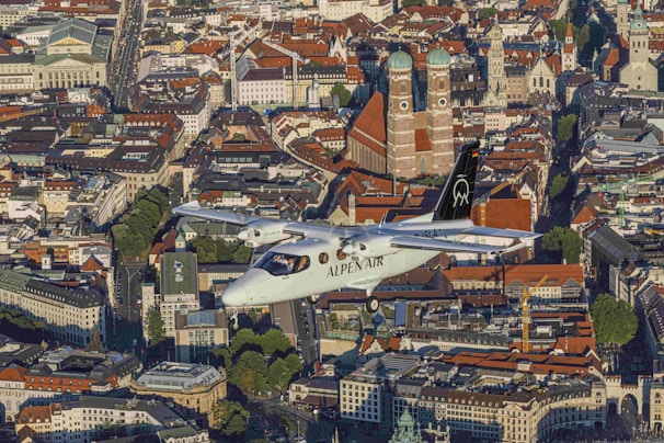An aircraft labeled 'Alpen Air' flies over a densely packed urban area characterized by historical architecture. Prominent buildings with red-tiled roofs stand out, including notable cathedrals with large domed towers. The city streets are lined with trees and various styles of architectural structures, reflecting a blend of modern and traditional designs.
