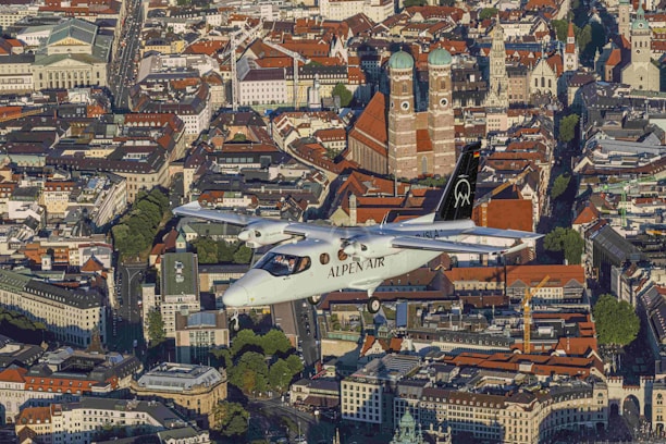 An aircraft labeled 'Alpen Air' flies over a densely packed urban area characterized by historical architecture. Prominent buildings with red-tiled roofs stand out, including notable cathedrals with large domed towers. The city streets are lined with trees and various styles of architectural structures, reflecting a blend of modern and traditional designs.