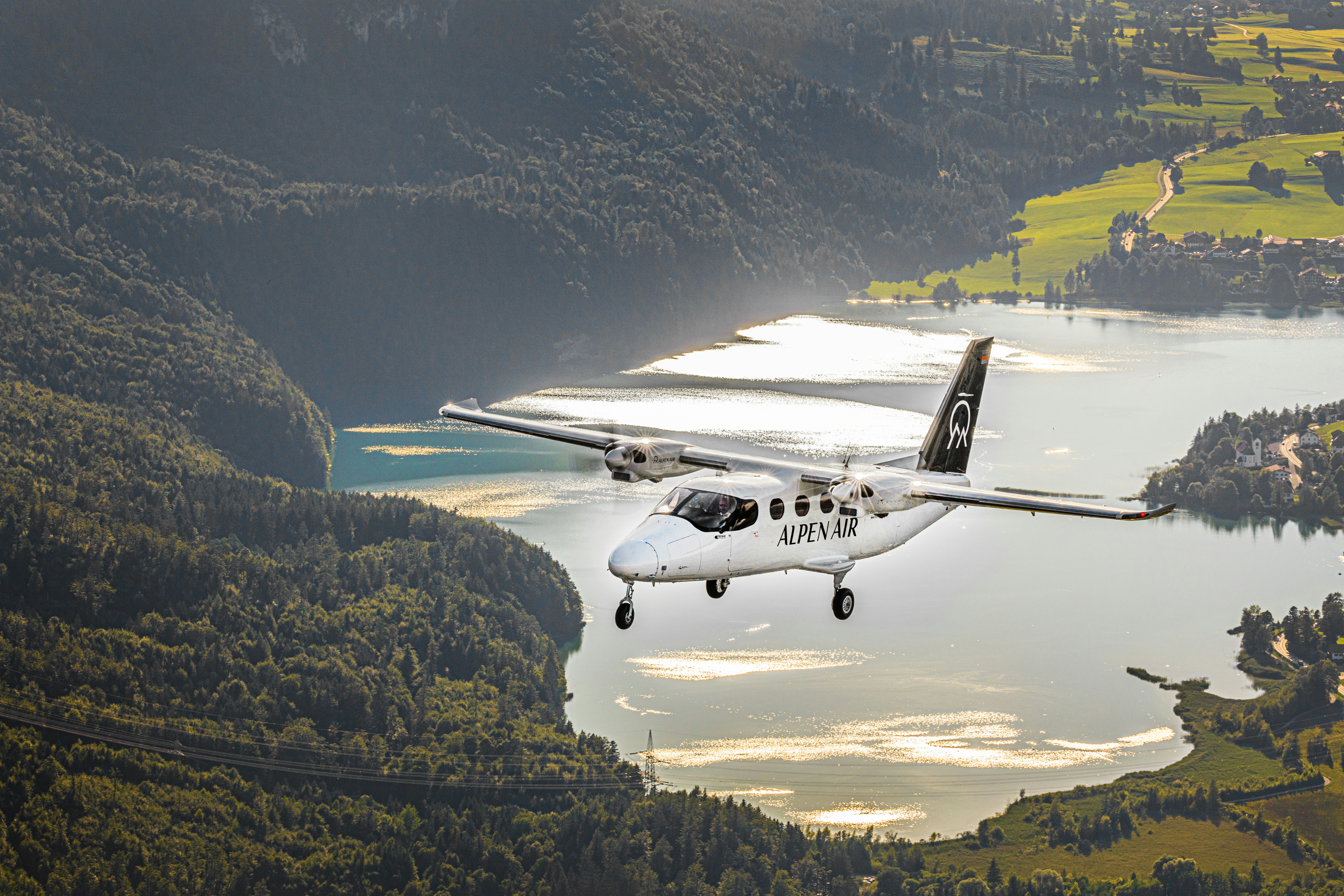 a small plane flying over a large body of water, The Alpen Air´s Tecnam P2012 Traveller aircraft above the Bavarian landscape with lakes, forests and meadow fields on a sunny day. Two pilots can be seen in the cockpit.