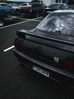 Several vehicles are parked in an outdoor parking lot. In the foreground, there is a black sports car with a GT-R emblem on the back and a license plate reading PSH384. The background shows more cars, including a blue car on a trailer and a pickup truck.