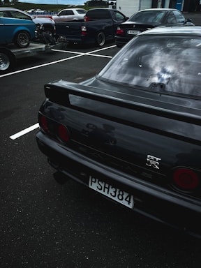 Several vehicles are parked in an outdoor parking lot. In the foreground, there is a black sports car with a GT-R emblem on the back and a license plate reading PSH384. The background shows more cars, including a blue car on a trailer and a pickup truck.